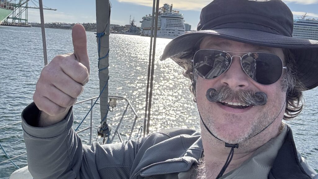 Andrew Mudd, on his boat in LA Harbor, wearing a big dumb hat and sunglasses, giving a thumbs up. Two cruise ships are in the background.