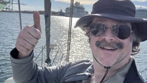 Andrew Mudd, on his boat in LA Harbor, wearing a big dumb hat and sunglasses, giving a thumbs up. Two cruise ships are in the background.
